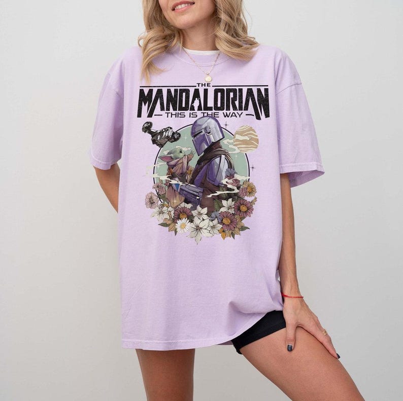 May include: A light purple t-shirt featuring a graphic from "The Mandalorian." The design includes the text "THE MANDALORIAN - THIS IS THE WAY" above an illustration of the Mandalorian and Grogu surrounded by flowers and a planet.