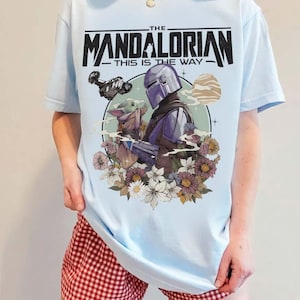 May include: Light blue t-shirt featuring a graphic of The Mandalorian and Grogu surrounded by flowers. The text "THE MANDALORIAN - THIS IS THE WAY" is printed across the top in black. The shirt is a short-sleeved crew neck.