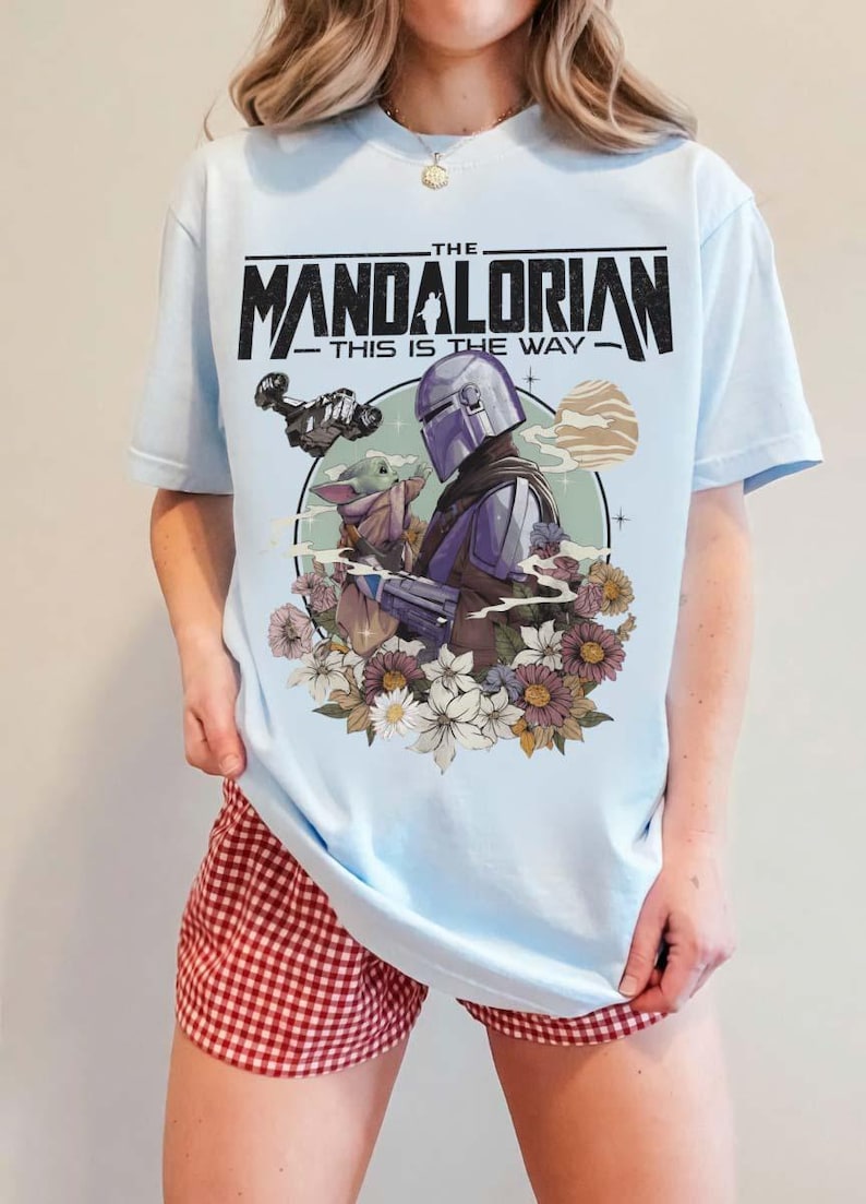 May include: Light blue t-shirt featuring a graphic of The Mandalorian and Grogu surrounded by flowers. The text "THE MANDALORIAN - THIS IS THE WAY" is printed across the top in black. The shirt is a short-sleeved crew neck.