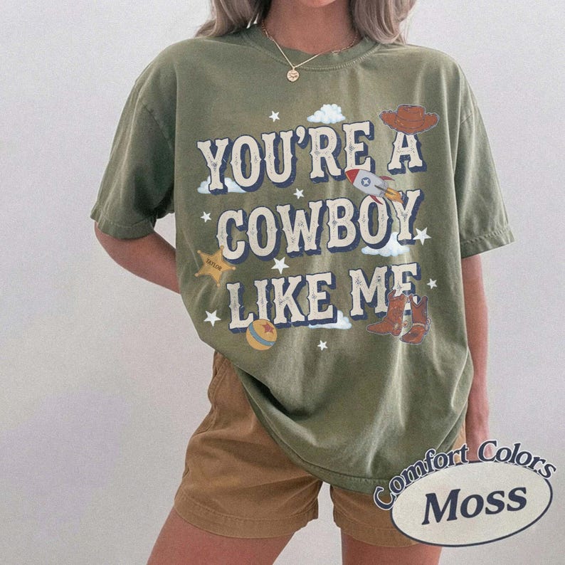 以下が含まれることがあります： オリーブグリーンの半袖Tシャツ。かすれた白文字で「YOU'RE A COWBOY LIKE ME」とプリントされています。ロケット、カウボーイハット、星、ボール、ブーツのイラストが描かれ、小さな白い星が散りばめられたデザインです。