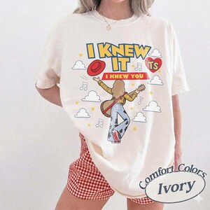 May include: An ivory short-sleeve t-shirt featuring a graphic print with the text I KNEW IT I KNEW YOU. The design includes a person with long hair playing a guitar, a red hat, a red heart with TS, yellow stars, and white clouds on a light background.