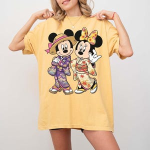 Puede incluir: Camiseta amarilla con una ilustraci&oacute;n de dibujos animados de Mickey y Minnie Mouse con trajes japoneses. Mickey lleva un sombrero de paja y un kimono morado, Minnie un lazo y un kimono con estampados florales. El dise&ntilde;o est&aacute; centrado en la camiseta.