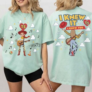 Puede incluir: Una camiseta verde claro con un gr&aacute;fico de un personaje vaquero tocando la guitarra en la parte delantera, rodeado de nubes, notas musicales y un coraz&oacute;n con las letras TS. La parte trasera muestra el texto I KNEW IT y I KNEW YOU con un personaje.