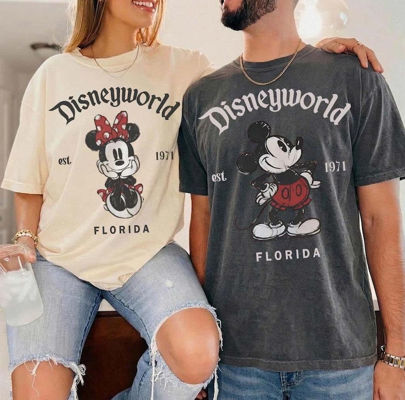 May include: Two people wearing matching t-shirts. One shirt is cream-colored with a Minnie Mouse graphic and the text "Disneyworld" and "Florida". The other shirt is gray with a Mickey Mouse graphic and the same text. Both shirts have "est. 1971".