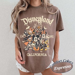 May include: Espresso-colored t-shirt featuring a Disneyland graphic with cartoon characters in skeleton costumes. The text includes "Disneyland," "Est. 1955," and "California." The shirt is a Comfort Colors brand.