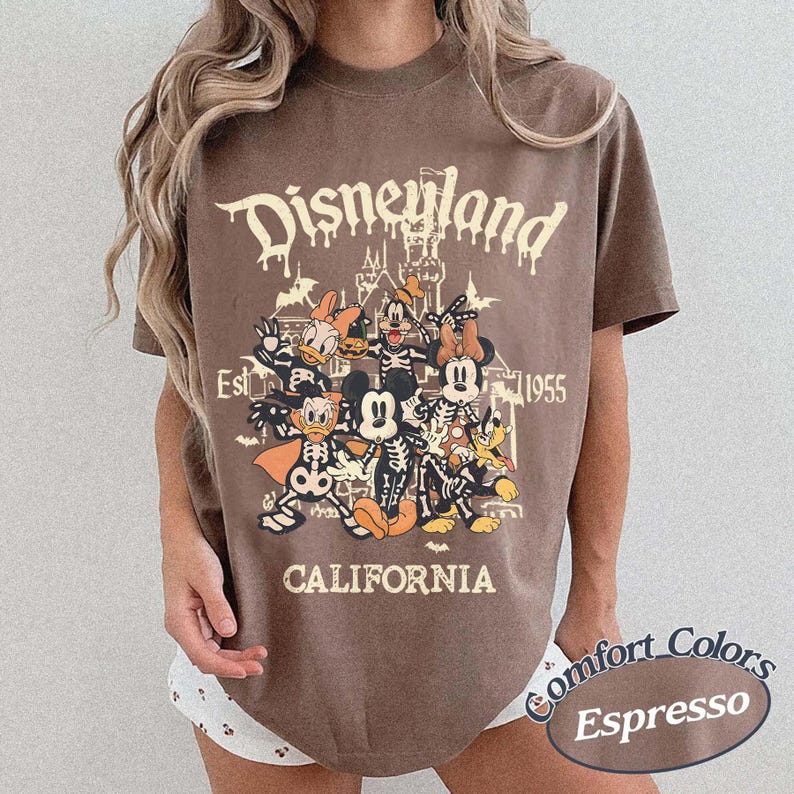 May include: Espresso-colored t-shirt featuring a Disneyland graphic with cartoon characters in skeleton costumes. The text includes "Disneyland," "Est. 1955," and "California." The shirt is a Comfort Colors brand.