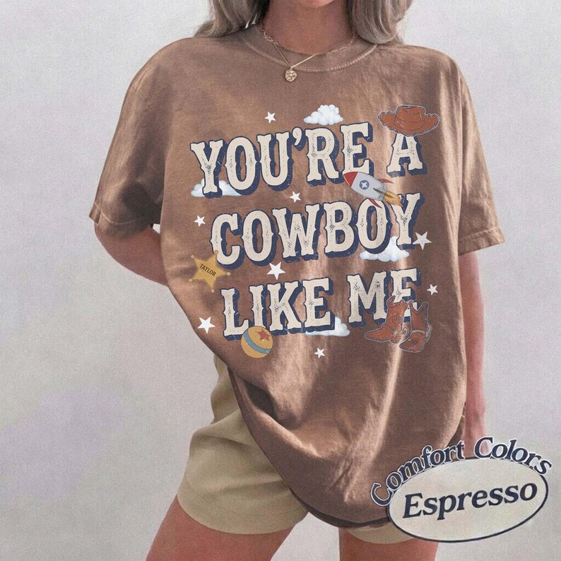以下が含まれることがあります： 「YOU'RE A COWBOY LIKE ME」という文字がかすれたフォントでプリントされた茶色の半袖Tシャツ。グラフィックには、ロケット、カウボーイハット、星型のバッジ、ボール、カウボーイブーツのイラストが描かれており、小さな白い星が散りばめられています。