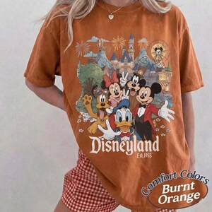 Pu&ograve; includere: Una t-shirt color arancio bruciato con una stampa grafica di Topolino, Minnie, Paperino, Pippo e Pluto davanti a un castello. Il testo recita Disneyland Est. 1955. La maglietta ha maniche corte e un girocollo con una vestibilit&agrave; rilassata.