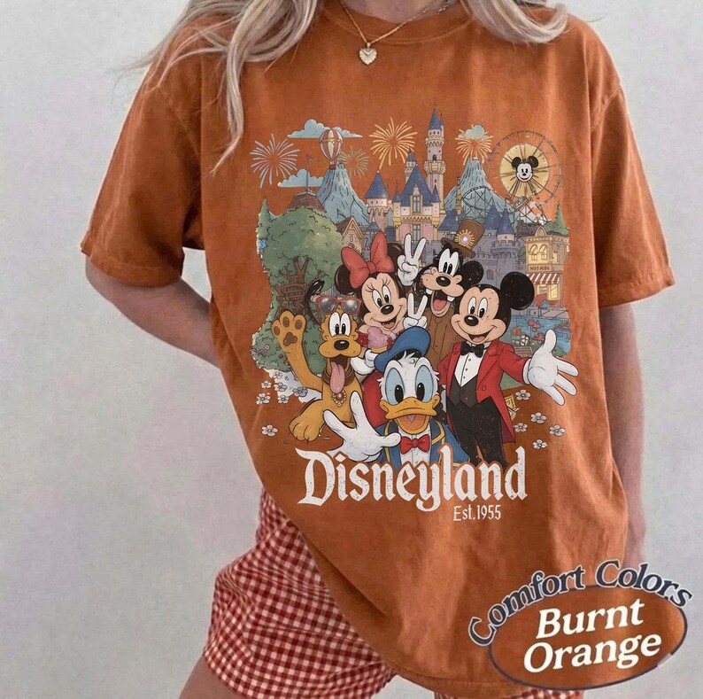 Pu&ograve; includere: Una t-shirt color arancio bruciato con una stampa grafica di Topolino, Minnie, Paperino, Pippo e Pluto davanti a un castello. Il testo recita Disneyland Est. 1955. La maglietta ha maniche corte e un girocollo con una vestibilit&agrave; rilassata.