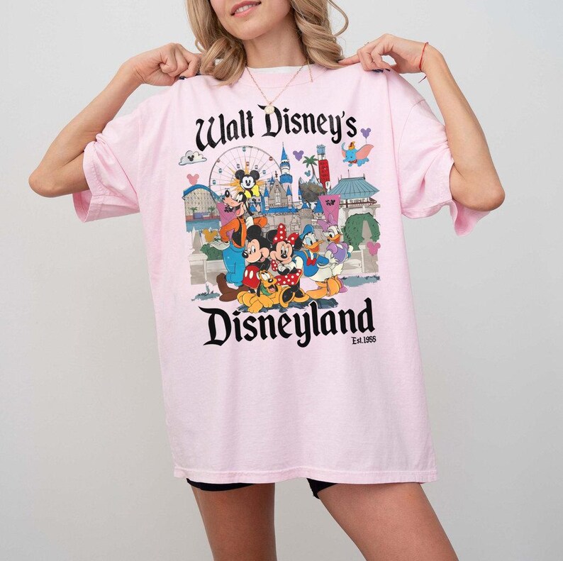 Puede incluir: Camiseta rosa claro con un estampado colorido de personajes y atracciones de Disneyland. El texto "Walt Disney's Disneyland" est&aacute; impreso en la parte superior e inferior. La camiseta tiene mangas cortas y un corte holgado.