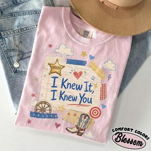 Op de afbeelding: Een lichtroze t-shirt met een afbeelding van een gelinieerde notitieblokpagina met de tekst "I Knew It, I Knew You" in het blauw. Het ontwerp bevat een gouden sheriffster, een cowboylaars, een wagenwiel, wolken, sterren, hartjes en tape.