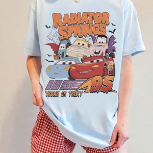 May include: Light blue t-shirt featuring a Halloween-themed graphic of characters from the movie Cars, with the text "Radiator Springs Trunk or Treat 95". The design includes bats and a spiderweb.