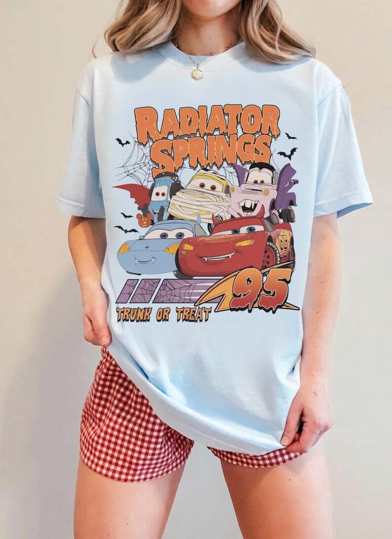 May include: Light blue t-shirt featuring a Halloween-themed graphic of characters from the movie Cars, with the text "Radiator Springs Trunk or Treat 95". The design includes bats and a spiderweb.