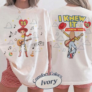 Puede incluir: Una camiseta color marfil con un gr&aacute;fico de una vaquera tocando la guitarra en la parte delantera y trasera. El frente muestra al personaje con notas musicales, nubes y un coraz&oacute;n con 'TS'. La parte posterior muestra el texto 'I KNEW IT I KNEW YOU'.