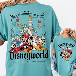 May include: Teal-colored t-shirt featuring a graphic of Disney characters in front of a castle, with the text "Disneyworld 1971." The design includes Mickey Mouse, Minnie Mouse, Donald Duck, Daisy Duck, Goofy, and Pluto. The shirt is a comfortable, casual style.