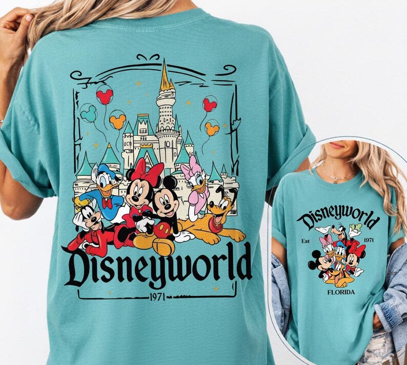 May include: Teal-colored t-shirt featuring a graphic of Disney characters in front of a castle, with the text "Disneyworld 1971." The design includes Mickey Mouse, Minnie Mouse, Donald Duck, Daisy Duck, Goofy, and Pluto. The shirt is a comfortable, casual style.