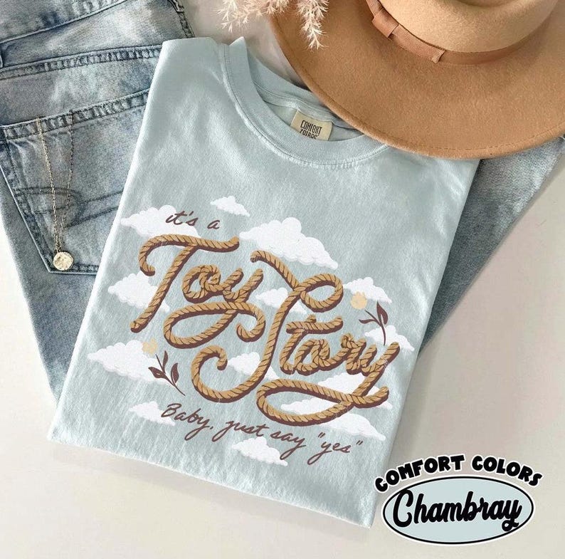 May include: A light blue t-shirt featuring a graphic of brown rope forming the text "It's a Toy Story" with white clouds and small floral accents. Below the rope text, the phrase "Baby, just say 'yes'" is printed in a dark brown script font.