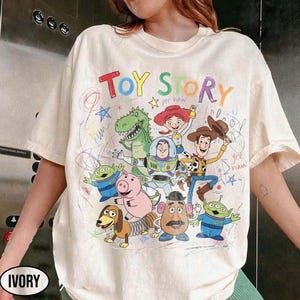 May include: Ivory-colored t-shirt featuring a colorful "Toy Story" graphic. The design includes cartoon characters like Woody, Buzz Lightyear, and Rex, with the words "Toy Story" in a playful font. The shirt has a crew neck and short sleeves.
