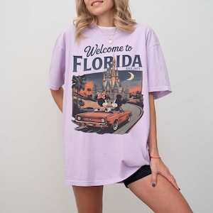 May include: A light purple t-shirt with the words "Welcome to Florida" and a graphic of Mickey and Minnie Mouse in a convertible driving towards a castle. The graphic also includes palm trees and the text "EST. 1971".