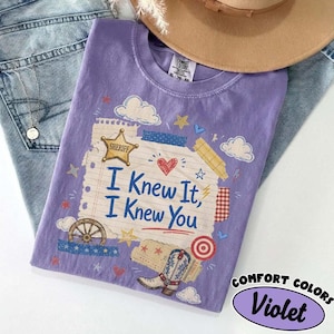 Op de afbeelding: Een violet t-shirt met een centrale afbeelding van gelinieerd notitiepapier met de tekst I Knew It, I Knew You. Het ontwerp bevat een gouden sheriffster, een rood hart, een wagenwiel, een cowboylaars en diverse kleine sterren en wolken.