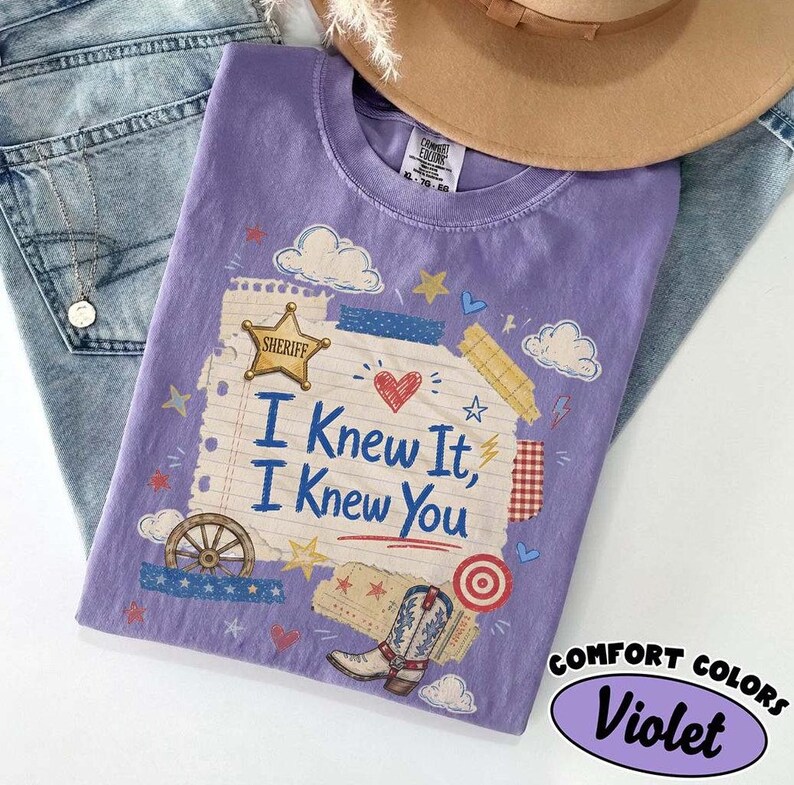 Op de afbeelding: Een violet t-shirt met een centrale afbeelding van gelinieerd notitiepapier met de tekst I Knew It, I Knew You. Het ontwerp bevat een gouden sheriffster, een rood hart, een wagenwiel, een cowboylaars en diverse kleine sterren en wolken.