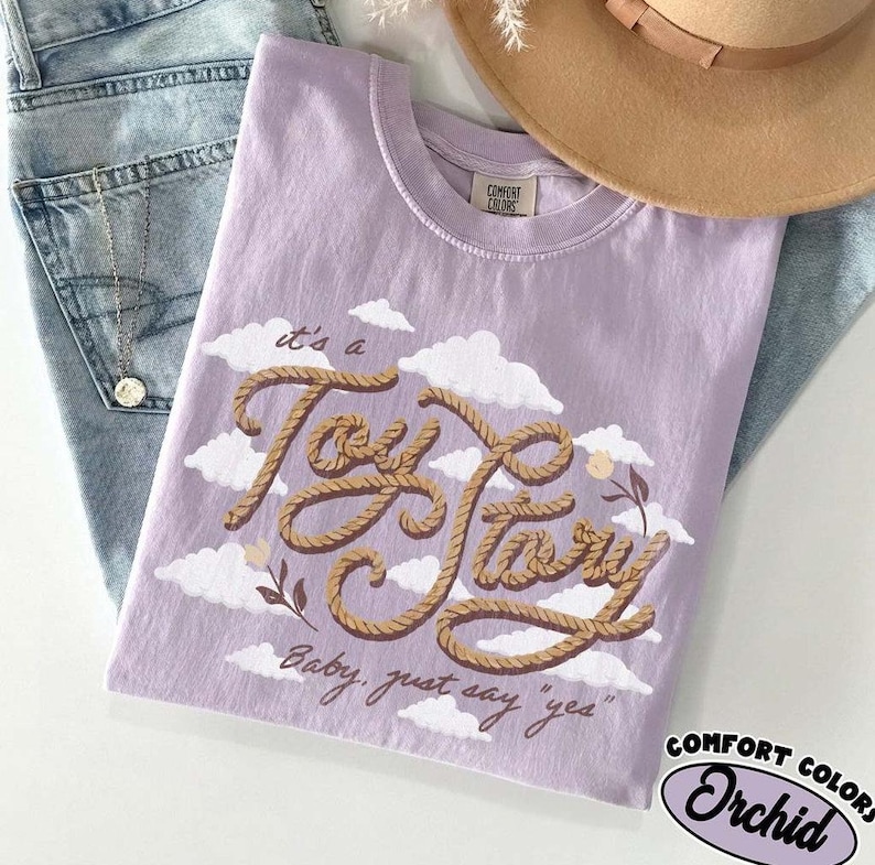 May include: A light purple t-shirt featuring a graphic of brown rope forming the words "it's a Toy Story" and the text "Baby, just say "yes"" below it. The design includes white clouds and small flowers on a light purple fabric background.