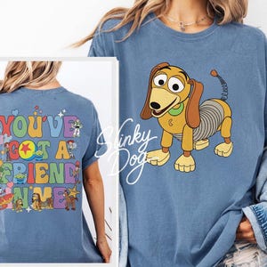May include: A blue t-shirt featuring a graphic of Slinky Dog on the front and a colorful design on the back with the text "YOU'VE GOT A FRIEND IN ME" surrounded by various cartoon characters including Buzz Lightyear, Woody, and Rex.