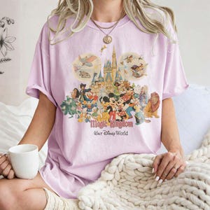 May include: A light purple t-shirt featuring a colorful graphic of Disney characters, including Mickey Mouse, Donald Duck, and Simba, in front of Cinderella's castle. The text "Magic Kingdom Walt Disney World" is below the graphic.
