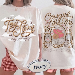 May include: An ivory t-shirt featuring a front graphic with rope-style text reading 'it's a Toy Story' and 'Baby, just say yes' amidst clouds. The back graphic displays 'Cowgirl like Me' and 'Takes One to Know One' with rope, a pink hat, and stars.