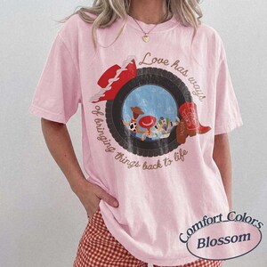 Może przedstawiać: Jasnor&oacute;żowy t-shirt z kr&oacute;tkim rękawem z grafiką czarnej opony otaczającej scenę z postaciami z kresk&oacute;wek. Tekst brzmi Love has ways of bringing things back to life. Na oponie umieszczono czerwony kowbojski kapelusz oraz czerwony kowbojski but.