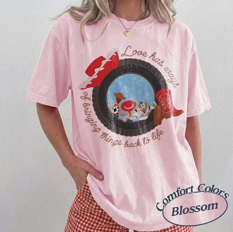 Może przedstawiać: Jasnor&oacute;żowy t-shirt z kr&oacute;tkim rękawem z grafiką czarnej opony otaczającej scenę z postaciami z kresk&oacute;wek. Tekst brzmi Love has ways of bringing things back to life. Na oponie umieszczono czerwony kowbojski kapelusz oraz czerwony kowbojski but.
