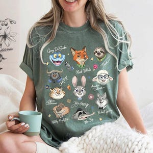 May include: Green t-shirt featuring cartoon characters from a movie. The shirt has illustrations of a frog, fox, sloth, sheep, bunny, and other characters. The names of the characters are written below each illustration. The shirt is a crew neck and has short sleeves.