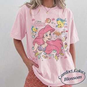 May include: Pink t-shirt featuring a graphic of Ariel from Disney's The Little Mermaid, with other characters and bubbles. The shirt has the text "Disney The Little Mermaid" and "Comfort Colors Blossom".