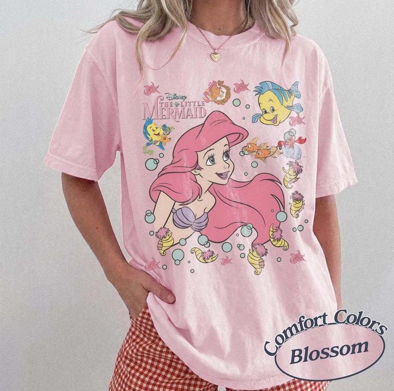 May include: Pink t-shirt featuring a graphic of Ariel from Disney's The Little Mermaid, with other characters and bubbles. The shirt has the text "Disney The Little Mermaid" and "Comfort Colors Blossom".
