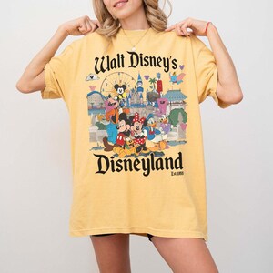 Puede incluir: Camiseta amarilla mostaza con un dise&ntilde;o de Disneyland. El dise&ntilde;o incluye el texto "Walt Disney's Disneyland" con varios personajes de Disney y puntos de referencia del parque. La camiseta tiene mangas cortas y un corte holgado.