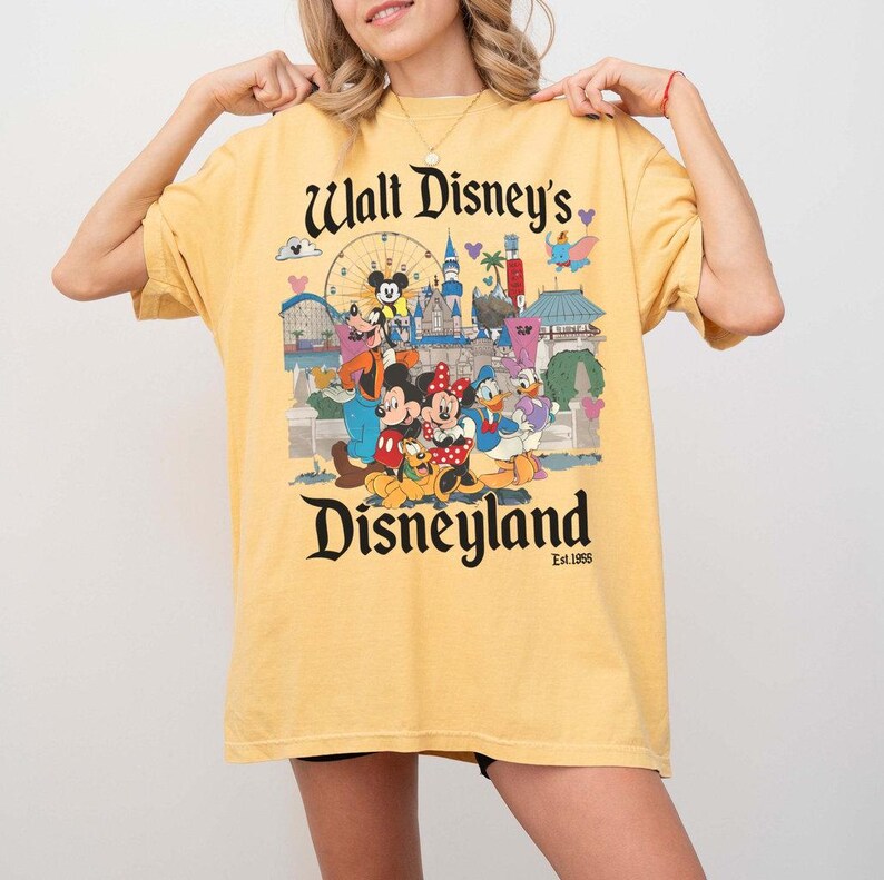 Puede incluir: Camiseta amarilla mostaza con un dise&ntilde;o de Disneyland. El dise&ntilde;o incluye el texto "Walt Disney's Disneyland" con varios personajes de Disney y puntos de referencia del parque. La camiseta tiene mangas cortas y un corte holgado.