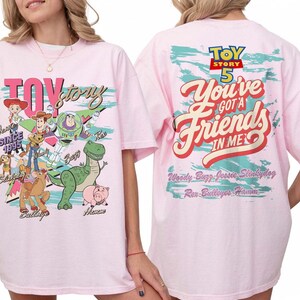 Pode incluir: Uma t-shirt rosa claro com gr&aacute;ficos de Toy Story. A frente mostra Woody, Buzz Lightyear, Jessie, Rex, Slinky Dog, Bullseye e Hamm com o texto Toy Story. As costas apresentam o texto Toy Story 5 You've Got A Friend In Me.