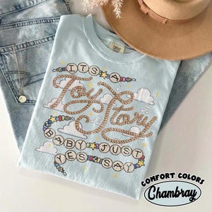 May include: A light blue t-shirt featuring a graphic design with the text "IT'S A Toy Story BABY JUST YES SAY" written in rope-style lettering and circular beads. The design includes white clouds, small stars, and musical note symbols.