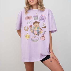 May include: A light purple t-shirt featuring a graphic print of a blonde cowgirl, a guitar, a horseshoe, a sheriff star labeled Swift Sheriff, a disco ball, a paper airplane, a cowboy boot, and a beaded necklace with the text Taylor's Story.