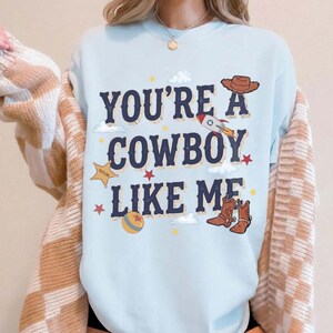 以下が含まれることがあります： ライトブルーのクルーネックトレーナー。ダークブルーの文字で「YOU'RE A COWBOY LIKE ME」とプリントされ、茶色のカウボーイハット、ロケット、黄色の保安官バッジ、カラフルなボール、茶色のカウボーイブーツのグラフィックイラストが周囲に配置されています。