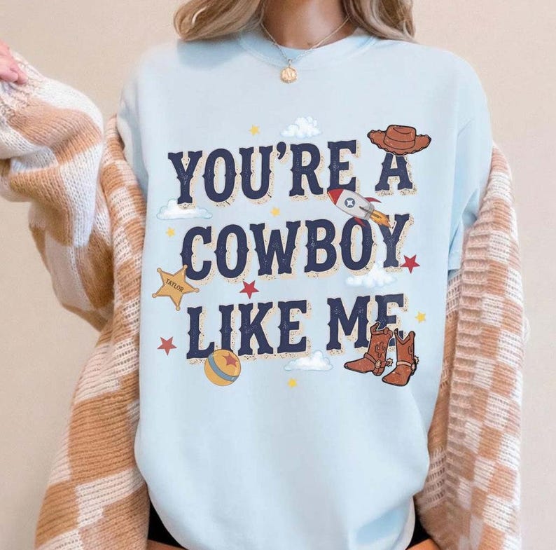 以下が含まれることがあります： ライトブルーのクルーネックトレーナー。ダークブルーの文字で「YOU'RE A COWBOY LIKE ME」とプリントされ、茶色のカウボーイハット、ロケット、黄色の保安官バッジ、カラフルなボール、茶色のカウボーイブーツのグラフィックイラストが周囲に配置されています。