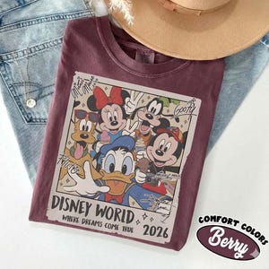 May include: A maroon t-shirt featuring a graphic of Disney characters, including Mickey Mouse, Minnie Mouse, Donald Duck, Goofy, and Pluto. The graphic reads "Disney World" and "2026". The shirt is paired with blue jeans and a tan hat.