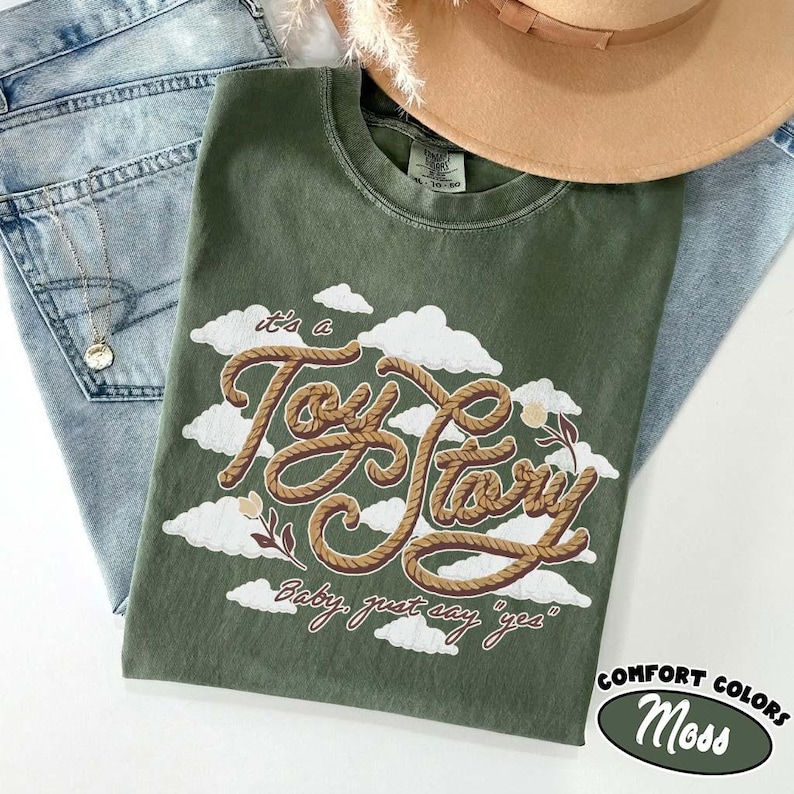May include: An olive green t-shirt featuring a graphic of white clouds and the text "it's a Toy Story" written in a brown rope-like font, with the additional text "Baby, just say "yes"" below it, accompanied by small floral illustrations.
