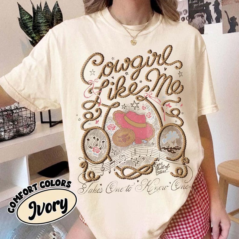 May include: An ivory short-sleeve t-shirt featuring a graphic print of a rope frame, a pink cowboy hat, a horseshoe, and musical notes. The text reads Cowgirl Like Me, Eyes full of Stars, and Takes One to Know One in a decorative script font.