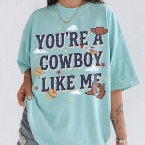 以下が含まれることがあります： 「YOU'RE A COWBOY LIKE ME」という紺色の文字がプリントされた水色のTシャツ。茶色のカウボーイハット、ロケット、黄色の保安官バッジ、カラフルなボール、拍車付きの茶色のカウボーイブーツのグラフィックイラストが描かれています。