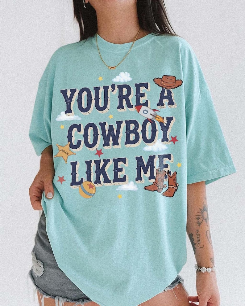 以下が含まれることがあります： 「YOU'RE A COWBOY LIKE ME」という紺色の文字がプリントされた水色のTシャツ。茶色のカウボーイハット、ロケット、黄色の保安官バッジ、カラフルなボール、拍車付きの茶色のカウボーイブーツのグラフィックイラストが描かれています。