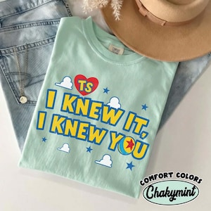 Puede incluir: Una camiseta de color verde menta claro con un coraz&oacute;n rojo con las letras TS, el texto I KNEW IT, I KNEW YOU en amarillo y un gr&aacute;fico de una pelota con una estrella roja, rodeado de peque&ntilde;as estrellas azules y nubes blancas en el frente.