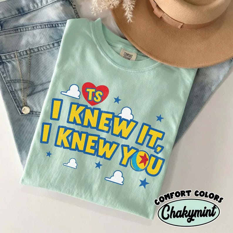 Puede incluir: Una camiseta de color verde menta claro con un coraz&oacute;n rojo con las letras TS, el texto I KNEW IT, I KNEW YOU en amarillo y un gr&aacute;fico de una pelota con una estrella roja, rodeado de peque&ntilde;as estrellas azules y nubes blancas en el frente.