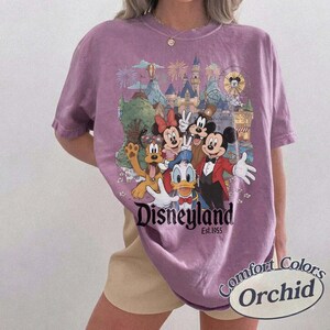 Pu&ograve; includere: Una t-shirt a maniche corte color orchidea con una stampa grafica di Topolino, Minnie, Paperino, Pippo e Pluto davanti a un castello. Il testo Disneyland Est. 1955 &egrave; stampato sotto i personaggi. La maglia ha un girocollo.