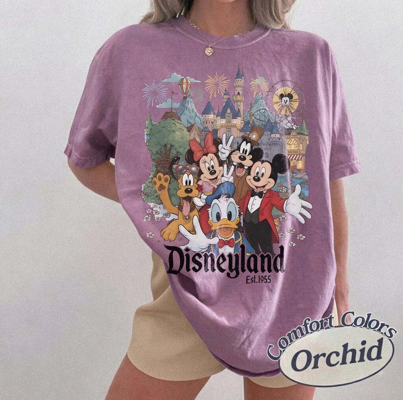 Pu&ograve; includere: Una t-shirt a maniche corte color orchidea con una stampa grafica di Topolino, Minnie, Paperino, Pippo e Pluto davanti a un castello. Il testo Disneyland Est. 1955 &egrave; stampato sotto i personaggi. La maglia ha un girocollo.
